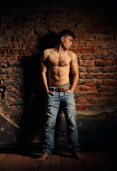 Un joven en jeans y con el torso desnudo de pie, apoyado contra una pared de ladrillo . — Foto de Stock