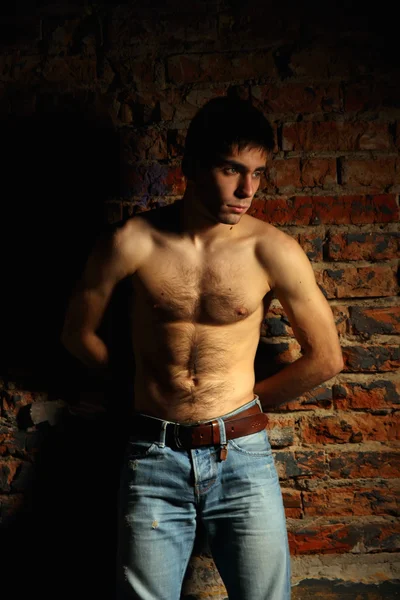 Un joven en jeans y con el torso desnudo de pie, apoyado contra una pared de ladrillo . — Foto de Stock