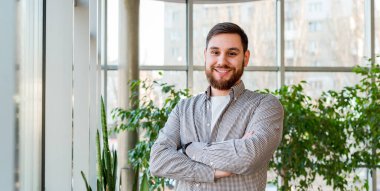 Evdeki seranın penceresinin yanında bitkilerle ya da modern ofislerle duran gülümseyen beyaz bir adam. Başarılı mutlu sakallı genç adamın portresi. Yakışıklı iş adamı. Uzun web pankartı