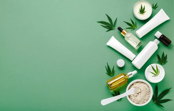 Beyaz model paketlerde kenevir cilt bakımı kozmetik seti. Nemlendirici krem, serum, losyon, CBD yağı, esaslı yağ keneviri yaprakları. Fotokopi alanı olan yeşil arkaplan üzerinde düz uzanır