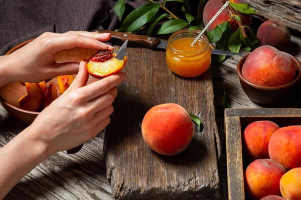 Woman hands cuts peaches for making recipe for jam, dessert, juice on rustic cutting board. Peaches whole fruits with leaves, peaches in halves, peach slices on wooden table. Dark mood.