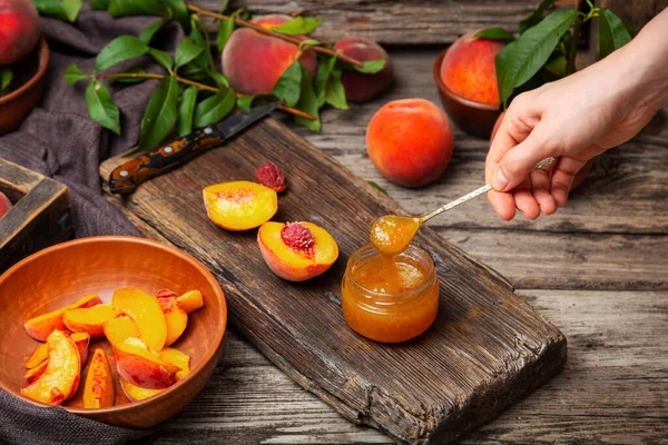 Peach jam spoon in female hand. Canned fruit jam glass jar with peach wedges and whole fruit. Peach jam on wooden table with ingredients. Process of making canned prescription jam.