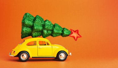 Sarı retro oyuncak araba çatıda şenlikli noel ağacı taşıyor. Arabanın çatısında yılbaşı ağacı. Kopyalama alanı yaratıcı minyatür Xmas ağacıyla turuncu renk arkaplanı. Uzun web pankartı.