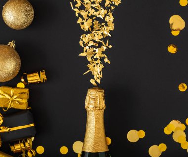 Altın şampanya şişesi altın pırıltılarını altın renkli Noel dekorları çerçevesinde saçar. Konfeti topları siyah arka planda hediyeler. Yılbaşı arifesinde düzlük çöktü. Karanlık arka plan kutlaması. Squere.