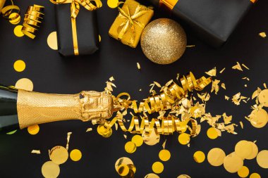 Altın şampanya şişesi altın pırıltılarını altın renkli Noel dekorları çerçevesinde saçar. Konfeti topları siyah arka planda hediyeler. Yeni yıl arifesi kutlamaları karanlık arka planda.