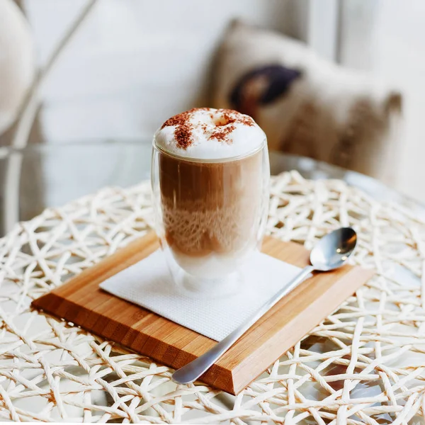 Hot latte or Cappuccino coffee on table in cafe or kitchen near the window.Morning coffee Latte with milk and chocolate on top in double walled glass on serving board with spoon. Square crop — Stock Photo, Image