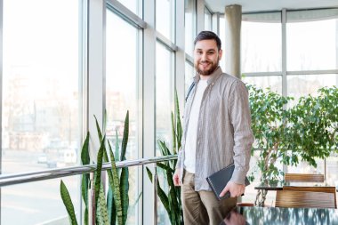 Sırt çantası, not defteri, üniversite kampüsünde ders kitapları olan bir öğrenci. Beyaz, genç, gülümseyen, sakallı, yakışıklı adam pencerenin yanında. Modern iş yerinde bitkilerle çalışan serbest çalışan biri.