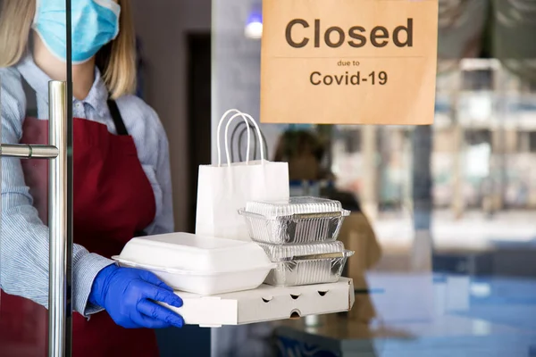 La camarera con máscara médica protectora y guantes trabaja con órdenes de comida para llevar. Camarero dando comida para llevar mientras está encerrado, cierre del coronavirus. Alimentos para entrega contra cartel cerrado debido a covid 19 — Foto de Stock