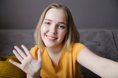 Mutlu kadın portresi el sallayarak web kamerası görüntüsünü selamlıyor. Genç ve güzel bir kadın oturma odasında kanepede otururken dizüstü bilgisayarla selam veriyor. Uzak çalışma çalışması, iletişim