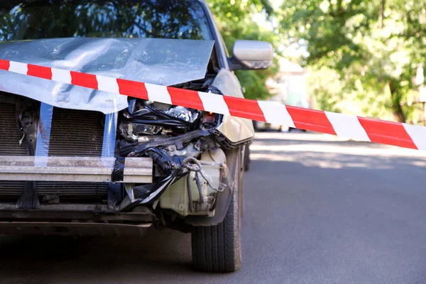 Destroyed car in car crash traffic accident on city road with red warning police tape. Smashed broken front auto headlight, dented hood without bumper on gray car accident. — Stock Photo, Image