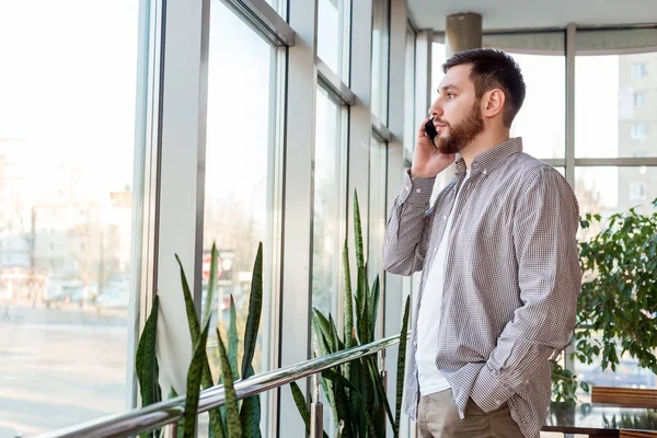 Smart beard Caucasian man is talking by smartphone. Businessman freelancer have Phone conversation in urban modern office in meeting room near window.