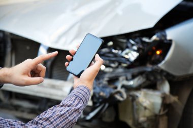 Adam araba kazasından sonra cep telefonu ekranını elinde tutuyor. İnternet uygulamasından araba kazası yerine sigorta şirketini arıyorum. Araba enkazının önünde akıllı telefon.