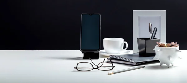 stock image Home Office workspace. Desktop with smart phone template blank, notepads pens office suppliers cup of coffee drink plant. White work desk table on black background. Long web banner with copy space.