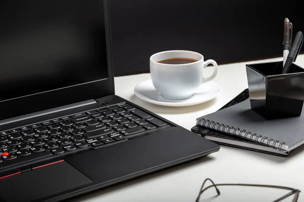 Черный ноутбук на белом столе черный фон. Очки, чашка кофе, канцтовары. Домашнее рабочее пространство для работы или учебы в интерьере офиса — стоковое фото