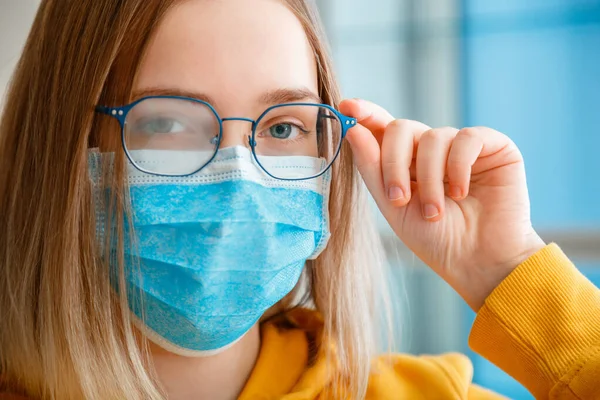 Óculos enevoados a usar em mulheres jovens. Retrato de perto. Menina adolescente em azul máscara protetora médica e óculos limpa embaçado nevoeiro misted óculos com espaço de cópia. Novo normal. — Fotografia de Stock