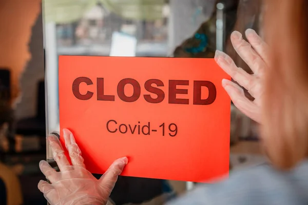 Firma Cerrado covid 19 bloqueo en la puerta de entrada de la tienda como nuevo cierre normal. Mujer con guantes protectores de máscara médica cuelga letrero cerrado en la puerta principal de la cafetería. Lockdown coronavirus covid 19 — Foto de Stock