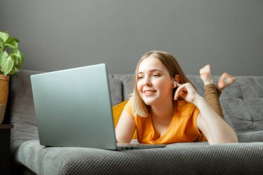 Happy, rahatla. Genç bayan, kulaklıklı dizüstü bilgisayarla kanepede uzanıyor. Uzaktan çalışma, çevrimiçi çalışma, online alışveriş, film izleme ya da dizüstü bilgisayardaki video görüşmesi. Genç kız oturma odasının iç tarafında dinleniyor..