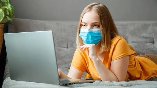 Woman in medical protective mask work using laptop at Home office lies on couch. Teen girl in mask Online learning education via laptop covid 19 lockdown time. Remote work Coronavirus pandemic banner.