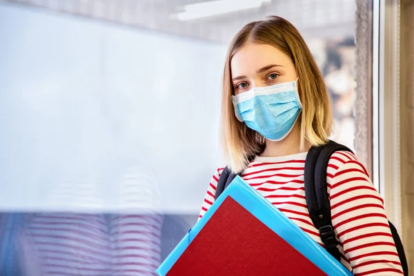 Girl student in protective medical mask. Portrait of blonde female student near window at university during coronavirus covid lockdown with copy space.