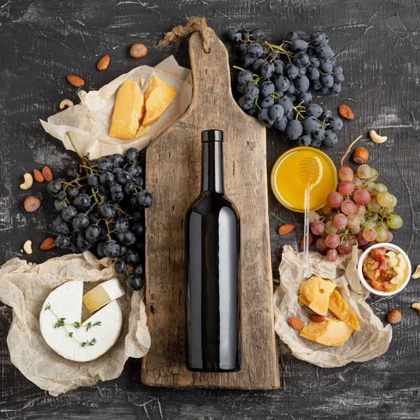 Wine drink mockup. Red wine bottle on vintage cutting wooden board. Frame made from Gastronomy of different cheeses grapes honey nuts. Restaurant dinner, wine tasting on dark concrete background.