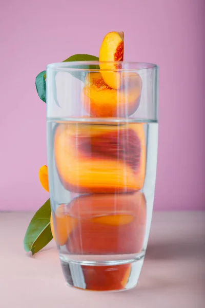 Flying fresh ripe peach in slices, whole fruits with green leaves. Peach fruit levitation on pink color background in frame of water, fruit behind glass of water. creative minimalistic concept peach