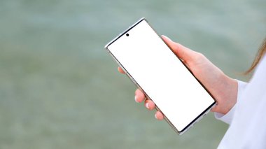 Beyaz model akıllı telefon, kadınların elinde. Kadın internette arama yapmak için cep telefonu, dışarıda uygulama kullanıyor. Deniz suyu yüzeyinde akıllı telefonlu kadın elleri. Kopyala uzayı Uzun web pankartı.