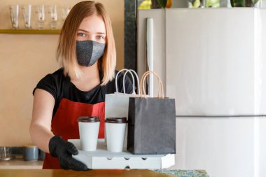 Mutfak çalışanı portresi çevrimiçi siparişleri eldiven ve maskeyle veriyor. Paket yemek kağıdı maketi. Yemek poşeti, pizza, içecek paketi paket servis kantinine. Temassız yemek teslimatı. Kapatma kodu 19.