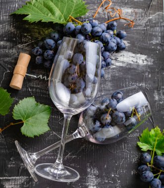 Siyah taş masada üzüm dolu şarap kadehleri. Koyu kırsal beton arka planda yaprakları ve sarmaşıkları olan üzüm demetleri. Şarap kadehi kompozisyonu ve üzüm taklit edilmiş taze kırmızı şarap. kare.