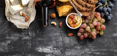 Yiyecek abur cuburları farklı peynirler, üzümler, koyu beton arka planda ballı şarap ekmeği içerir. Uzun web afişi. Şarap ve peynir kompozisyonu. Akdeniz Mutfağı gastronomi aperatifi Kırmızı şarap şişesi.