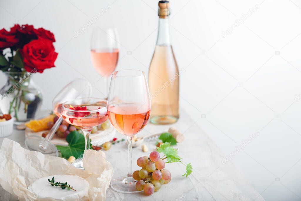 Rose wine in different wineglasses, bottle on white table with grapes cheese, snacks bouquet of flowers. modern still life Rose Wine composition on light grey concrete background with copy space.