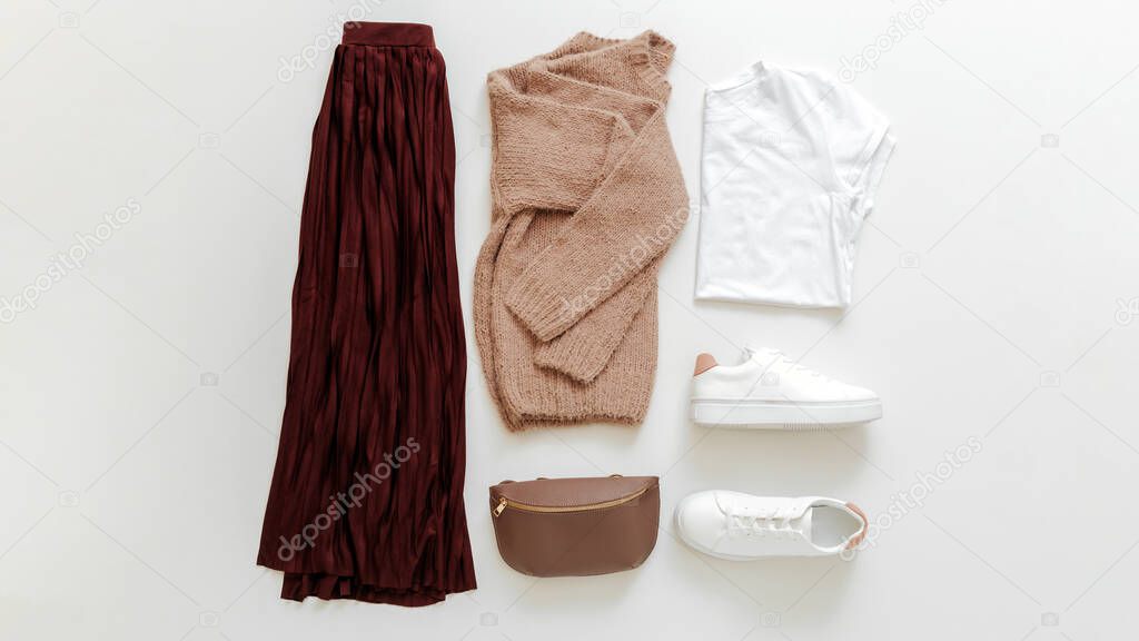 Female spring look autumn outfit burgundy skirt beige sweater white shoes sneakers bag white basic tshirt on white background flat lay. Folded clothes women fashion urban basic outfit. Long Web banner