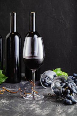 Siyah taş masada üzümlü şarap kadehi kompozisyonu. Kırmızı şarap şişeleri, yapraklı üzüm demetleri ve koyu kırsal beton arka planda sarmaşıklar. Kırmızı şaraplı şarap bardağı. Dikey.