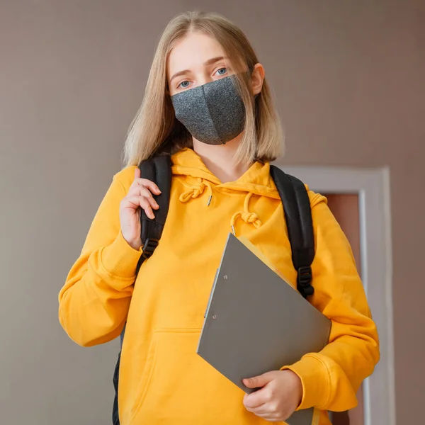 Schoolgirl in mask with backpack and notepad. Young woman student in protective medical mask. Portrait of blonde female student Girl at university interior during coronavirus covid 19 lockdow. Square.