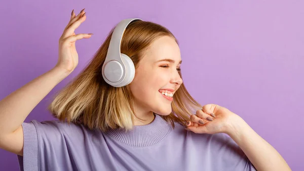 Happy smiling young woman dancing in headphones with flying blonde hair hairstyle. Teen girl enjoy listen music moving in wireless headphones isolated over purple color background. Long web banner.