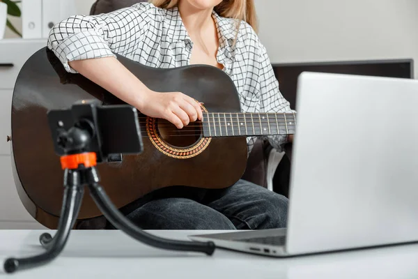 Young woman plays acoustic guitar at home for online audience on laptop and smartphone. Online classes course guitar training music e education during lockdown. Online musical guitar performance