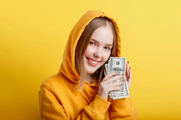 Emotional happy teenage blonde girl in hood win money cash holding dollars in hands isolated on color yellow background. Portrait young excited smiling woman with stack of money banknotes. — Stock Photo, Image