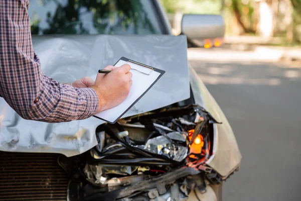 Male insurance agent with auto insurance blank against Destroyed car in car crash traffic accident on road. Smashed broken front auto headlight on car accident. Auto life and health insurance.