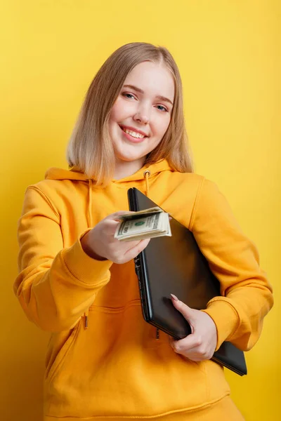 Belle jeune femme souriante pigiste avec ordinateur portable prendre billets d'argent dollars. IT travailleur à distance indépendant à distance via ordinateur portable isolé sur fond de couleur jaune. — Photo