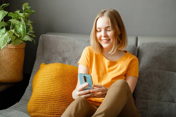 Smiling woman using smartphone at home. Teen blonde girl hipster search chatting on phone at room on couch. Young woman use mobile app for chat online communication at home interior — Stock Photo, Image