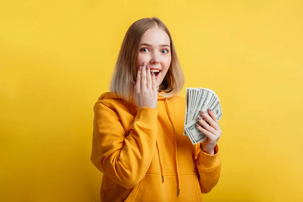 Šťastná nadšená bohatá mladá žena vyhrává peníze. Lady držet Dolar zásobník překvapený raduje z vítězství v loterii. Dospívající dívka zakryl její otevřená ústa s rukou překvapení izolované přes žluté barvy pozadí — Stock fotografie