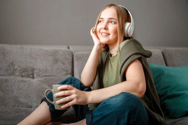 Glückliche junge Frau, die auf dem Sofa sitzt und Kopfhörer mit einer Tasse Tee trägt. Frau oder Teenie-Mädchen in Ruhe, Glückseligkeit genießen Musik auf der Couch zu Hause im Wohnzimmer hören — Stockfoto