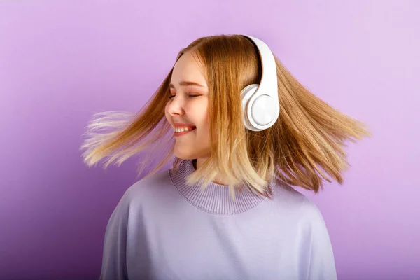 Attractive smiling woman dancing in headphones with flying blonde hair hairstyle. Teenager girl enjoy listen music moving in wireless headphones isolated over purple color background.
