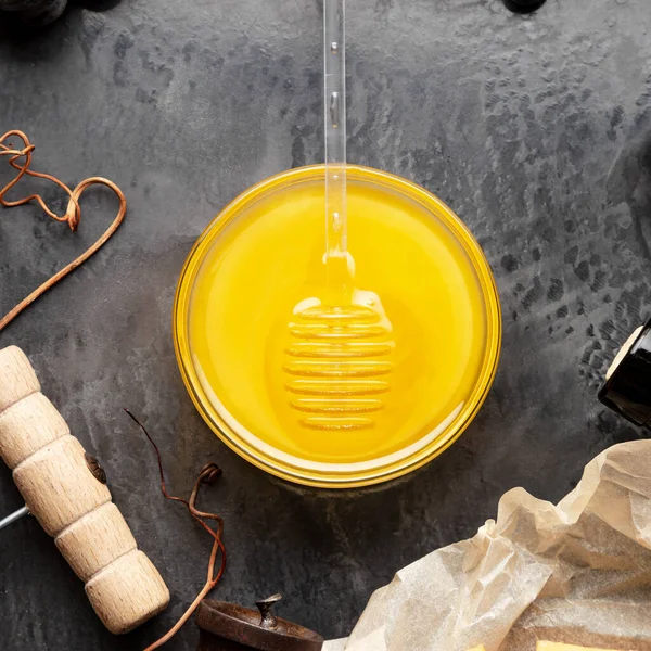 Honey in glass jar with honey spoon on dark stone board with wine cheese corkscrew in Black juicy grapes frame. Delicious yellow bee honey in snack plate on black table. Top view. Square photo — Stock Photo, Image