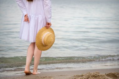 Deniz kıyısında elinde hasır şapkayla tanınmayacak kadar mutlu bir kadın. Beyaz plaj elbiseli, ince bacaklı kadın silueti deniz sahilinde gün batımında dinleniyor.