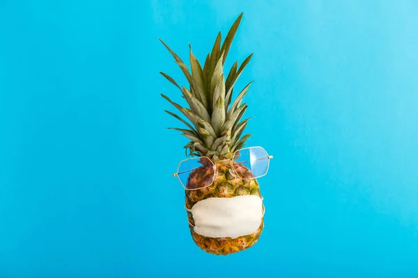 Cara divertida piña en gafas de sol con estilo máscara médica en color azul fondo de verano. Concepto de viaje piña en máscara protectora en el bloqueo del coronavirus. — Foto de Stock