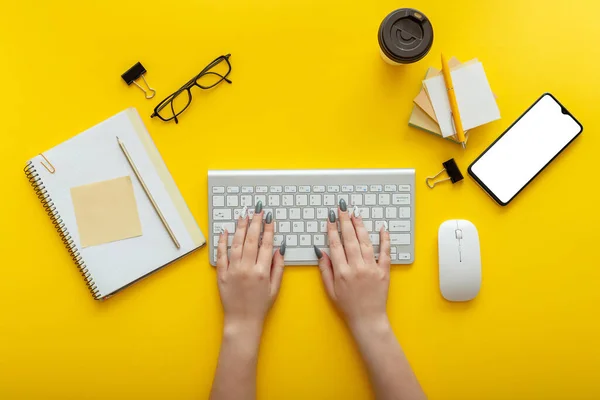 Woman typing on computer keyboard at workplace on color yellow background. Office desktop workspace with female hands, cup of coffee smartphone mockup office supliers, glasses. Flat lay Top view
