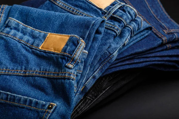 Denim jean pants folded in stack with empty brown tag mockup label. Casual wear blue jeans stacked in pile on black background. — Stock Photo, Image