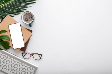 Akıllı telefon ekranı hazırlar, klavye kahve bardağı bardakları beyaz çalışma masasında yeşil bitki yaprakları. Modern ofis çalışma alanındaki cep telefonu. Üst görünüm düz çerçeve ve kopyalama alanı.