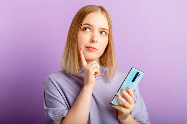 Young woman think doubt about question using smartphone. Teen girl curious dreaming, choose or decide look side on copy space. Emotion portrait isolated over purple color background — Stock Photo, Image