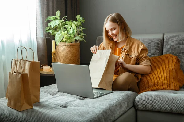 Happy young woman do unpacking online orders goods or food. Online Shopping, ordering delivery. Teenager girl relax on sofa considering purchases with laptop. Mock up paper bags